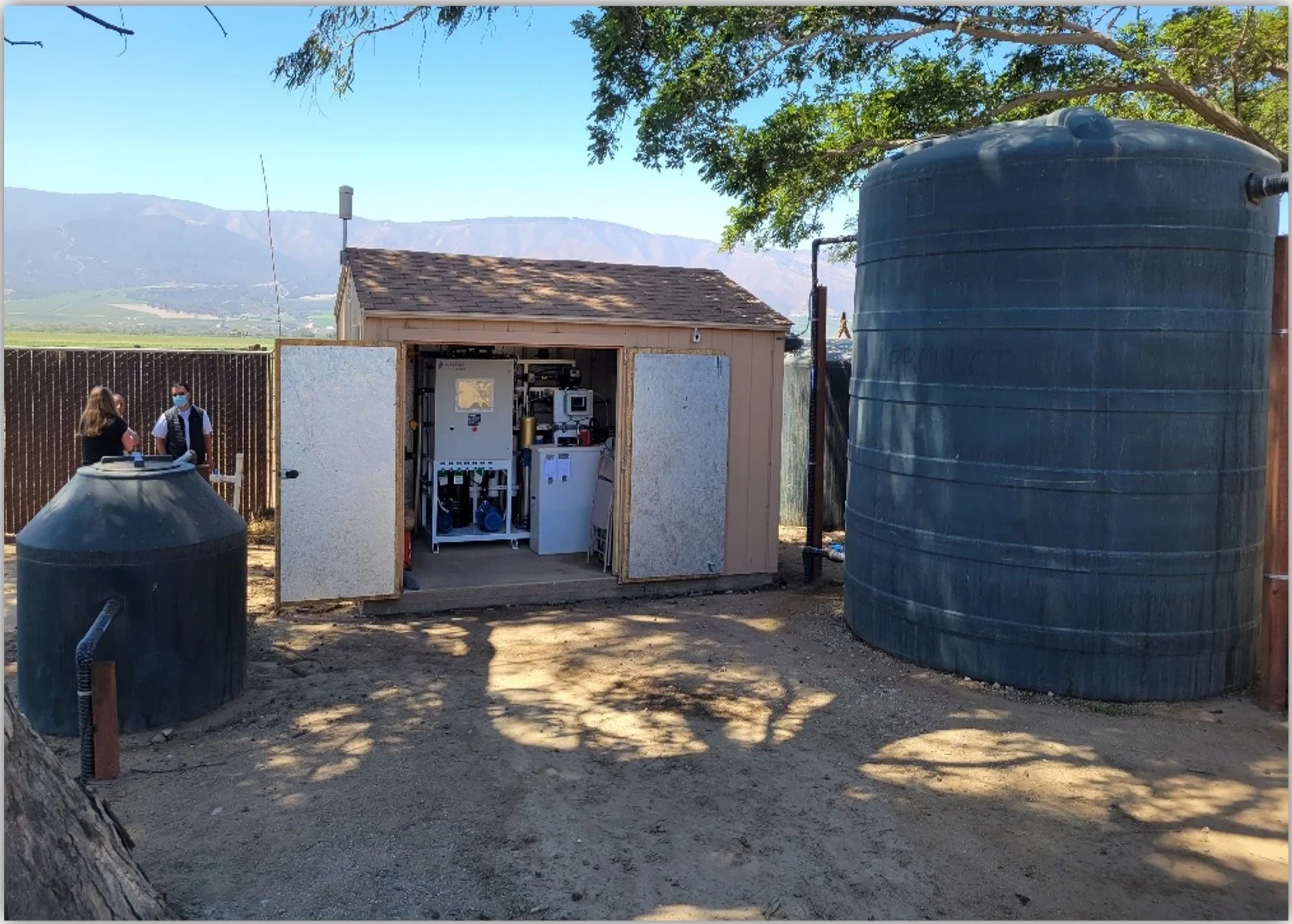 A small treatment building at a well, doors open, with water tanks on either side and mountains behind